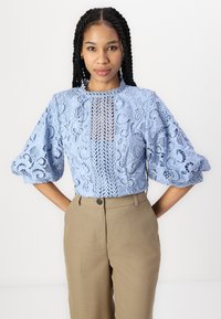 Lys blå blondebluse med intrikate blomsterdesign, puffy ermer og knappet halsåpning, kombinert med beige skræddersydde bukser.