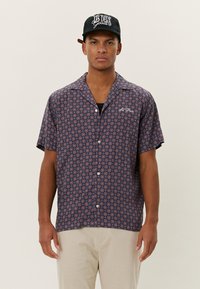Short-sleeve shirt in navy fabric with a circular red and white pattern, featuring a relaxed collar and front buttons. Worn with a black cap.