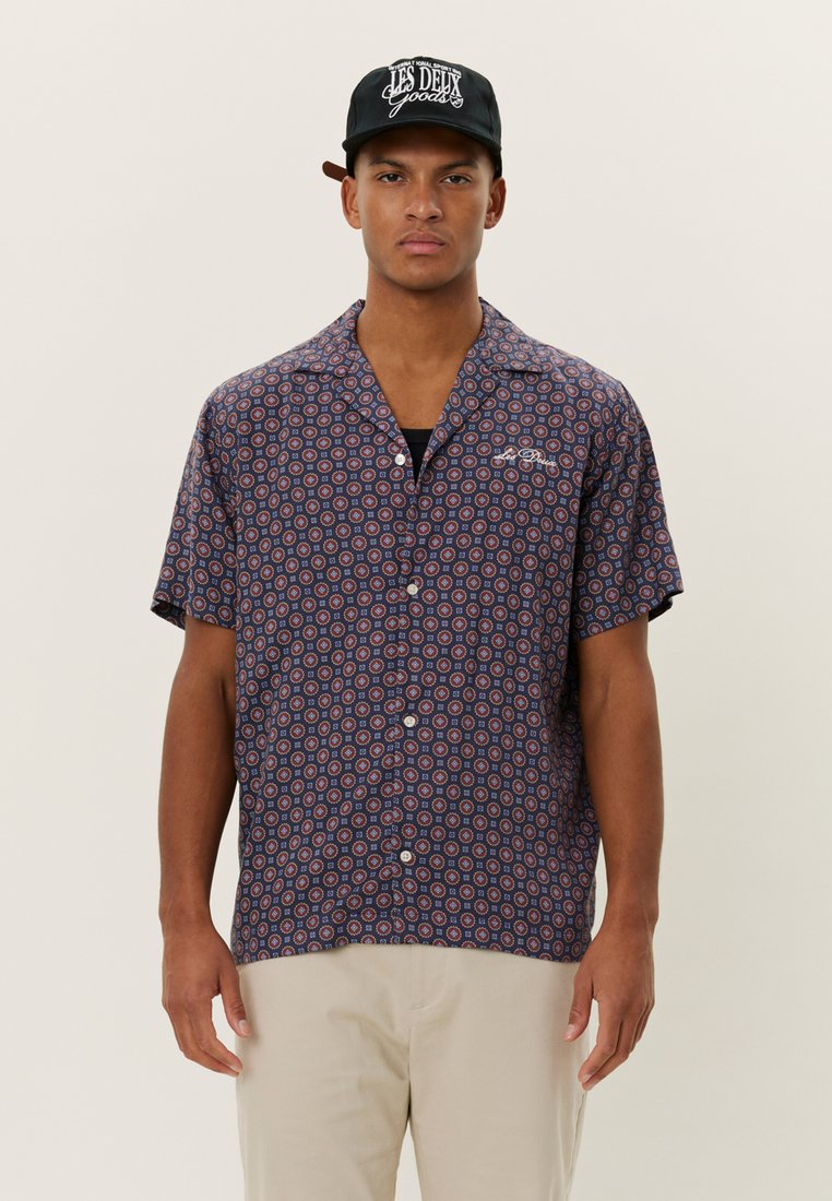 Short-sleeve shirt in navy fabric with a circular red and white pattern, featuring a relaxed collar and front buttons. Worn with a black cap.