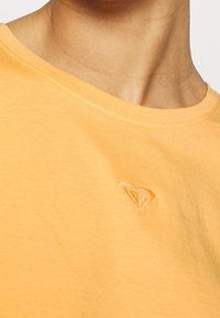 Detailní záběr na osobu oblečenou v žlutém tričku s kulatým výstřihem, na němž je na hrudi vyšitý malý logotyp ve tvaru srdce.