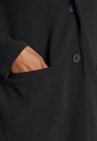 Czarny płaszcz wykonany z teksturowanej tkaniny, z widoczną kieszenią i zapięciem na guziki. Miękka i gładka struktura z prostymi detalami designu.