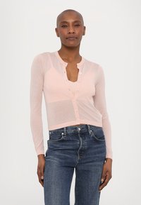 Lys rosa, gjennomsiktig cardigan med lange ermer, rund hals og knappelukking foran. Brukt over mørkeblå jeans.