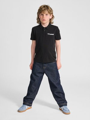 Ung dreng stående med benene fra hinanden, iført en sort polo med "hummel" logo, løse mørke jeans og blå sneakers med gummisåler.