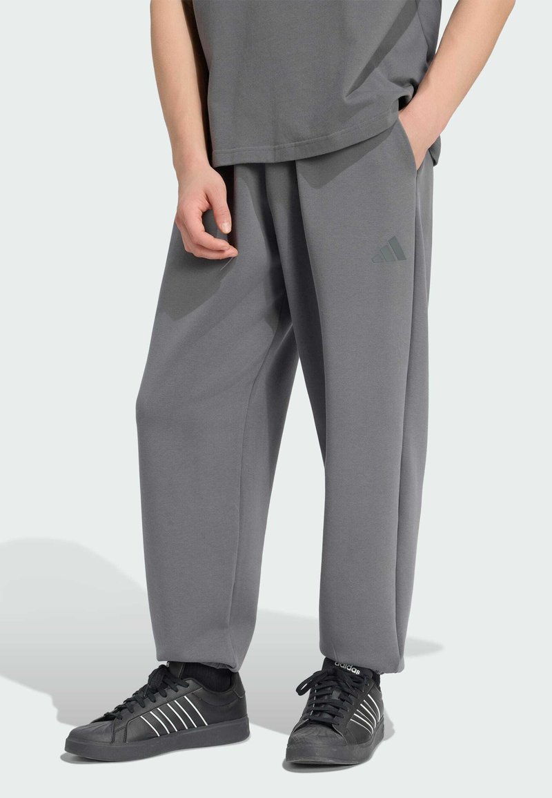 Personne portant un pantalon de survêtement gris ample Adidas et des baskets noires Adidas, debout avec une main dans la poche et l'autre détendue sur le côté.