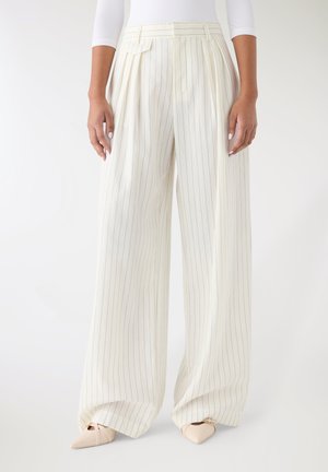 Pantaloni bianchi a gamba larga plissettati con righe verticali grigie. Presentano una vita alta e una texture liscia. Abbinati a ballerine beige con punta appuntita.