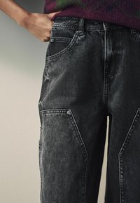 Personne portant un pantalon en jean noir avec de grandes coudières aux genoux et un pull bordeaux, main glissée dans la poche avant.