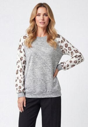Frau mit blondem Haar, die einen grauen Pullover mit weißen und braunen Leopardenmuster-Ärmeln und schwarze Hosen trägt, vor einem schlichten Hintergrund stehend.