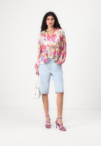 Blusa a maniche lunghe con fantasia floreale in rosa, giallo e blu, abbinata a bermuda in denim azzurro chiaro e sandali viola con cinturini.
