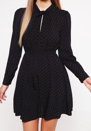 Czarna sukienka z długim rękawem z małymi brązowymi wzorami, z talią podkreśloną paskiem oraz dekoltem w szpic zawiązanym na kokardę, noszona przez osobę z długimi włosami.