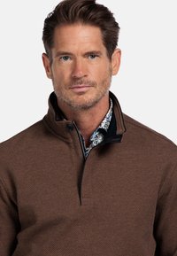 Man met kort bruin haar en lichte stoppelbaard, gekleed in een gestructureerde, bruine pullover over een bloempatronen shirt, staand met zijn gezicht naar voren tegen een witte achtergrond.