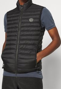 Schwarze gesteppte ärmellose Weste mit Reißverschluss vorne, Seitentaschen und Logo-Patch, getragen über einem blauen Kurzarmhemd und dunklen Hosen.