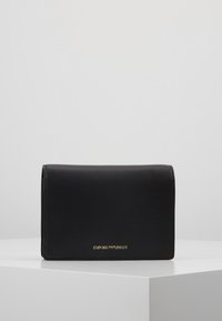 Černá kožená peněženka Emporio Armani s zlatým logem, umístěná na bílé geometrické podstavě proti jednoduchému šedému pozadí.