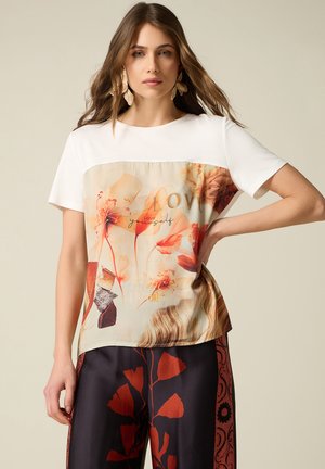 Kobieta z długimi włosami, ubrana w biały top z nadrukiem kwiatowym i napisem „Love yourself”, w połączeniu z czarnymi spodniami z wzorem czerwonych liści.