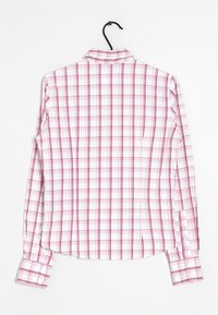 Camisa de botón de manga larga con una base blanca y un patrón de cuadros rosa; cuenta con un cuello clásico y puños cuadrados. Cuelga de un gancho negro.
