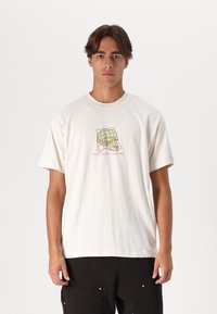 Camiseta de algodón color crema con un diseño gráfico que presenta un contorno de calavera y un patrón de cuadrícula en azul y rosa. Mangas cortas, cuello redondo.