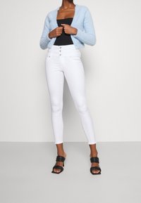 Cardigan de felpa azul claro, top negro ajustado, jeans skinny blancos de cintura alta y sandalias de tacón cuadrado negras. Diseño simple, sin patrones visibles.