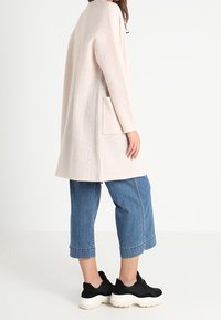 Hellrosa übergroßer Mantel mit langen Ärmeln und seitlicher Tasche; kombiniert mit kurzen blauen Jeans und schwarzen Plateau-Sneakern.