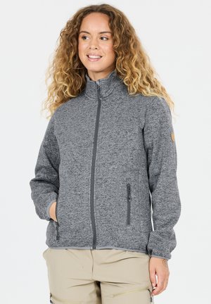 Nainen kiharat hiukset, harmaa vetoketjullinen fleecetakki ja beige housut, hymyilee ja katsoo sivulle. Kädet taskuissa.