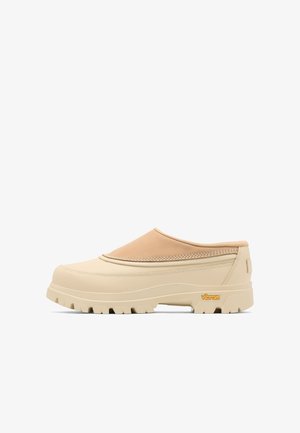 Scarpa beige senza lacci con suola Vibram spessa, dettagli cuciti e tomaia imbottita che copre il collo del piede, mostrata su sfondo bianco.