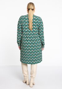 Robe verte à motifs avec des manches longues, un décolleté plongeant à l'arrière, et une coupe décontractée ; associée à des bottes beiges à talons épais montant jusqu'aux genoux.