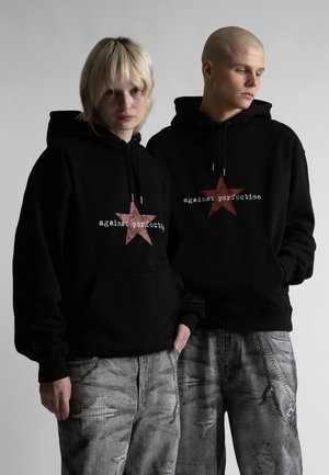 Dvaja ľudia v čiernych mikinách s kapucňou s červenými hviezdami a nápisom „against perfection“, v páre s opotrebovanými šedými džínami, stoja pred sivým pozadím.