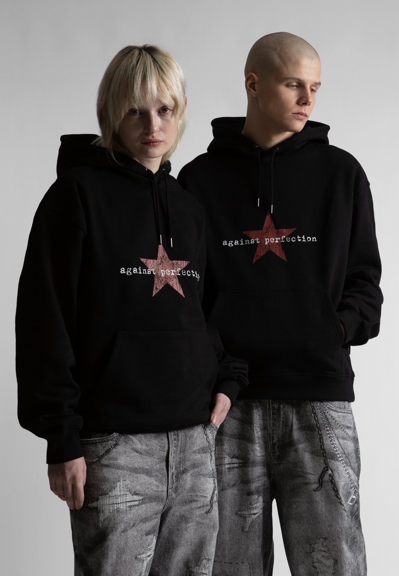 Δύο άτομα φορούν μαύρα φούτερ με κουκούλα με κόκκινα αστέρια και το κείμενο «κατά της τελειότητας», συνδυασμένα με γκρι τζιν με φθορές, στέκονται μπροστά σε γκρι φόντο.