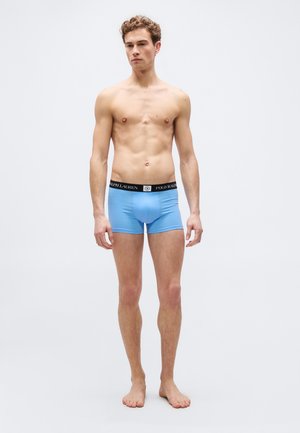 Junger Mann steht barfuß und trägt hellblaue Boxershorts mit schwarzem Polo Ralph Lauren Bund vor einfarbigem Hintergrund.