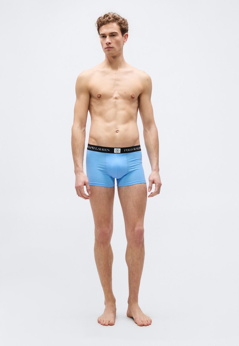 Νεαρός άνδρας όρθιος, ξυπόλητος, φοράει ανοιχτό  μπλε εσώρουχα boxer με μαύρη ζώνη Polo Ralph Lauren, σε απλό φόντο.