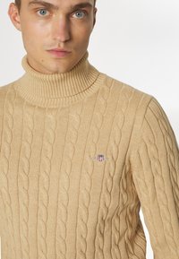 Jersey de cuello alto de punto beige con patrón de trenzas y cuello acanalado. Presenta un pequeño logo en el pecho, mostrando textura y diseño.