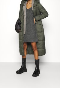 Zelená péřová bunda s kapucí a kostkovanou podšívkou, kombinovaná s šedými pletenými šaty a černými botami s robustní podrážkou. Texturovaný a vrstvený design.