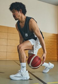 Juodas be rankovių sportinis marškinėliai, balti krepšinio šortai ir balti sportbačiai su juodais akcentais. Driblingas raudonu ir baltu krepšiniu ant sporto salės grindų.