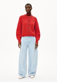 Persoon in een felrode gebuttonde cardigan, gestreepte coltrui, lichte wijde jeans en witte schoenen op een plain witte achtergrond.