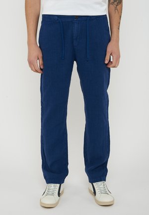 Uomo che indossa pantaloni blu navy con coulisse e sneakers bianche con striscia nera, in piedi davanti a uno sfondo semplice.