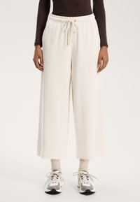 Széles lábú, krémszínű melegítőnadrág puha anyagból, rugalmas derékkal és zsinóros részletekkel. Pasztell zoknikkal és sportcipőkkel párosítva.