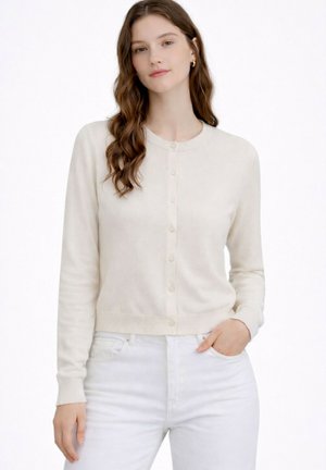 Jeune femme aux longs cheveux bruns portant un cardigan crème boutonné et un jean blanc, debout avec une main dans la poche.