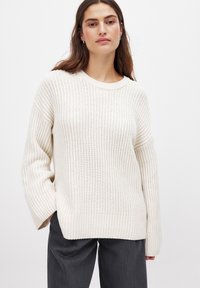 Heegeldatud jumpers heledas valges, ribateraga tekstuuriga, laiade varrukatega ja ümarate kaelustega, kombineeritud tumehallide pükstega.
