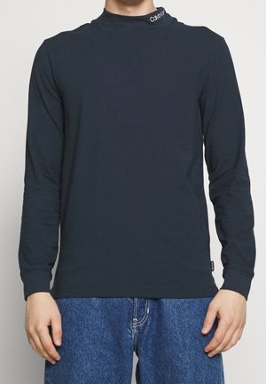 Mężczyzna ubrany w dopasowaną czarną koszulę z długim rękawem z logo Calvin Klein na kołnierzu oraz niebieskie dżinsy na jednolitym tle.