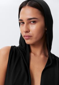 Black sleeveless hooded garment with a draped design, soft texture, and smooth finish, showcasing a relaxed fit around the neckline.