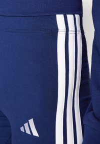 Tmavě modré sportovní kalhoty s přiléhavým střihem, zdobené třemi bílými svislými pruhy po straně a malým logem.