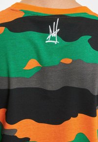 T-maica s camo uzorkom u zelenoj, narančastoj i crnoj boji. S okruglim izrezom, kratkim rukavima, s bijelim logotipom izvezenim na leđima. Pamuk.