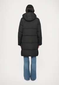 Melnā pūkaina jaka ar kapuci, rakstaina ar šūtu dizainu un vidēja garuma griezumu. Kombinēta ar gaiši ziliem zvanveida džinsiem.