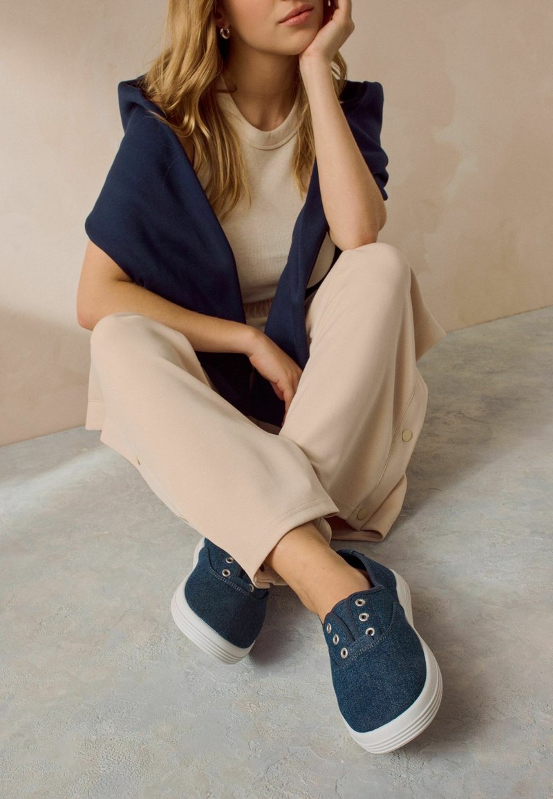 Blå canvas sneakers med hvide såler, der har snøreringe og en struktureret overflade, parret med beige afslappede bukser og en marineblå top.