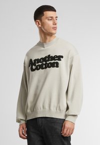 Világosszürke pamut pulóver, a mellrészen texturált fekete "Another Cotton" logóval, ribbelt mandzsetta és alj.