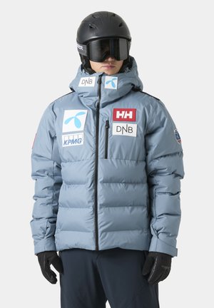 Person iført lyseblå polstret skijakke med sponsormærker, sort hjelm, skibriller og handsker, stående mod en hvid baggrund.