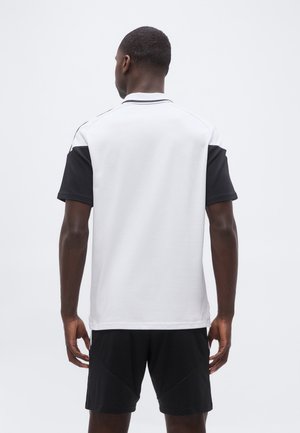 Homme portant une chemise blanche avec des manches courtes noires et un short noir, debout dos à la caméra devant un fond uni.