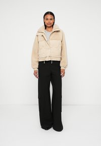 Giacca trapuntata beige con un esterno testurizzato e fuzzy, colletto alto e vita regolabile, indossata sopra una maglietta grigia e abbinata a pantaloni neri.
