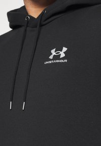 Sudadera negra con cordones, que presenta un logo de Under Armour bordado en plata en el pecho. Textura de tejido suave con un corte regular.