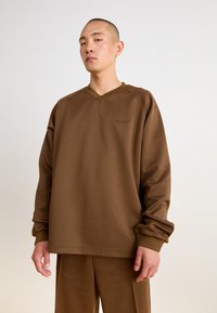 Sweatshirt marron à manches longues avec un col en V et des poignets côtelés, arborant un logo discret sur la poitrine. Associé à un pantalon marron assorti.