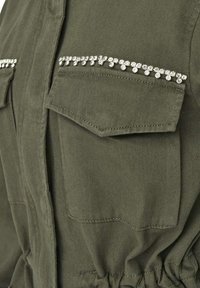 Veste utilitaire vert olive avec poches poitrine doubles ornées de strass, taille à cordon de serrage et tissu texturé.