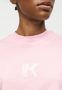 Κοντινό πλάνο ενός ατόμου που φοράει ανοιχτό ροζ πουκάμισο με ένα διακριτικό λευκό λογότυπο που περιλαμβάνει τα γράμματα "IK" και μια μικρή σιλουέτα.